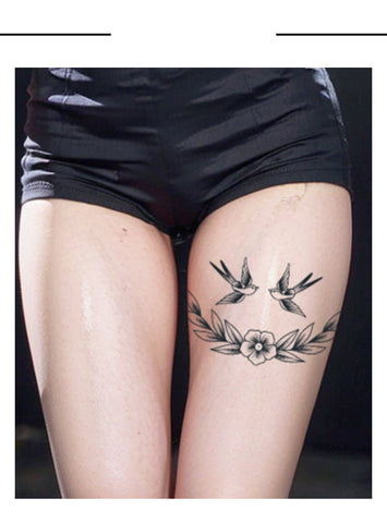 Tattoo design of two birds and a flower on a person's leg with black shorts.